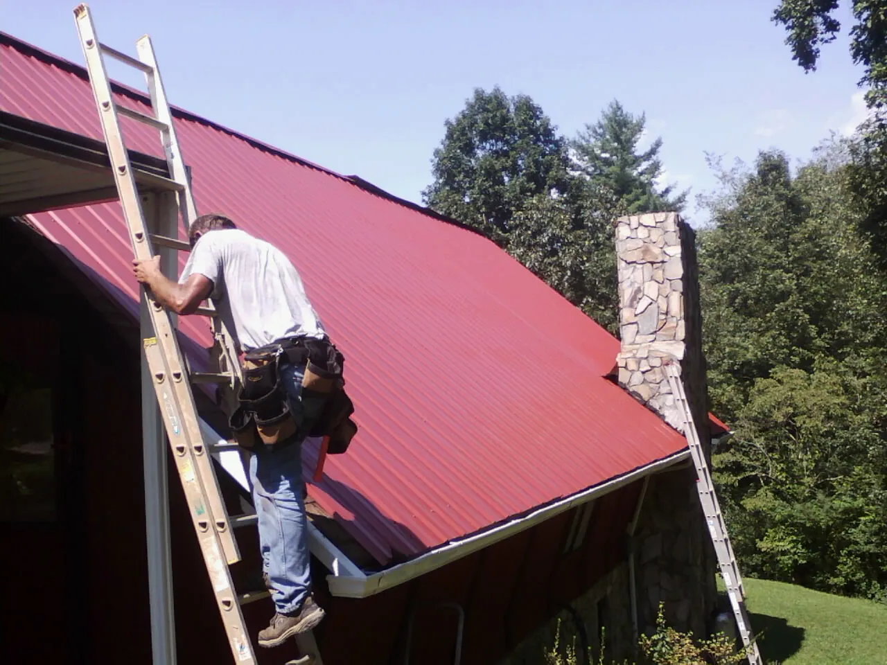 Roofer on a ladder beside a red metal roof and stone chimney — Soffit & Fascia Repair by Roofing Brothers in Lillian